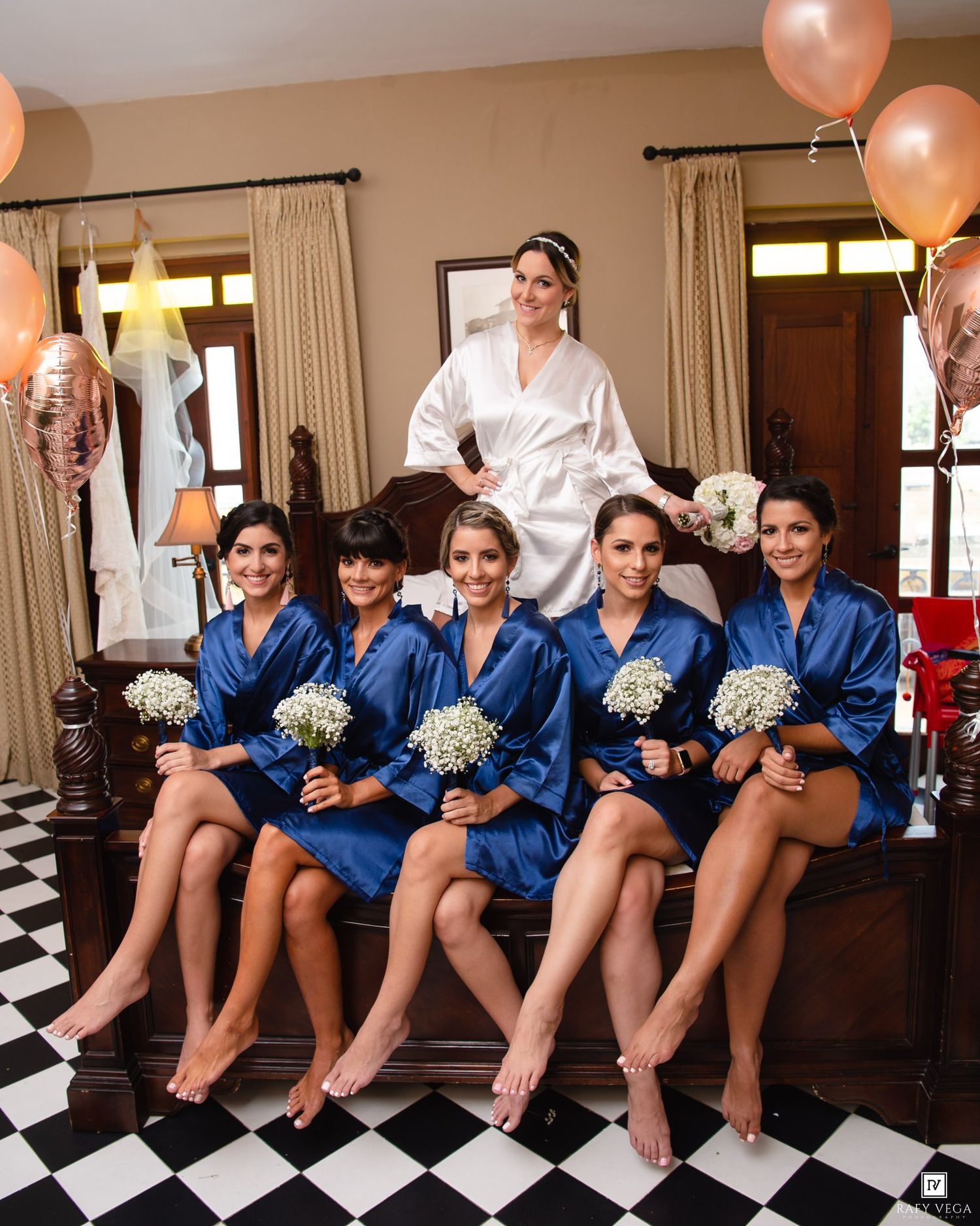 Bride in white robe stands behind four bridesmaids in blue satin robes holding bouquets on a bed in a decorated room.