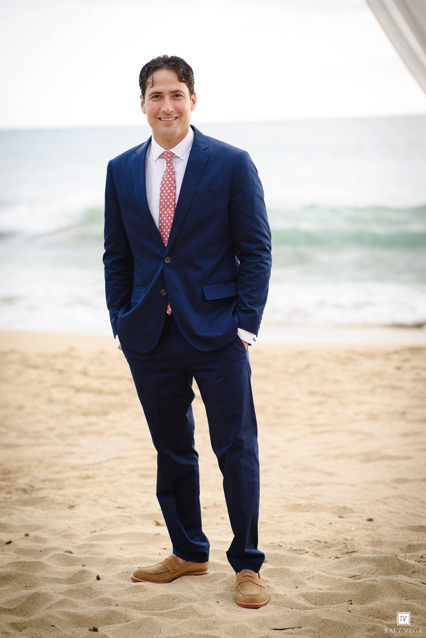 Man in navy blue suit with pink tie standing on sandy beach with ocean waves in background.