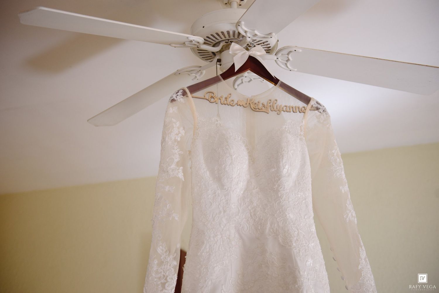 Elegant lace wedding dress hanging from a personalized bride hanger on a white ceiling fan in a bridal suite.