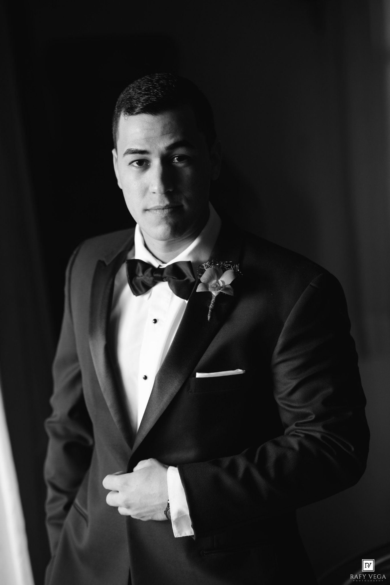 Elegant groom in a black tuxedo with bow tie and boutonniere poses for a formal wedding portrait.