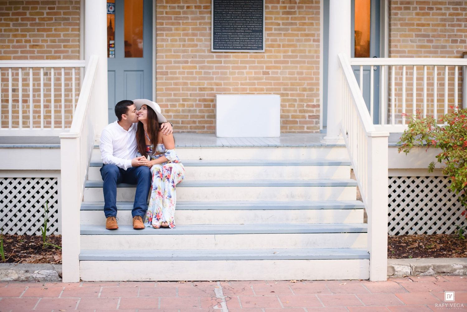 Couple sitting on white porch steps of brick building, sharing a romantic moment in casual and floral attire.