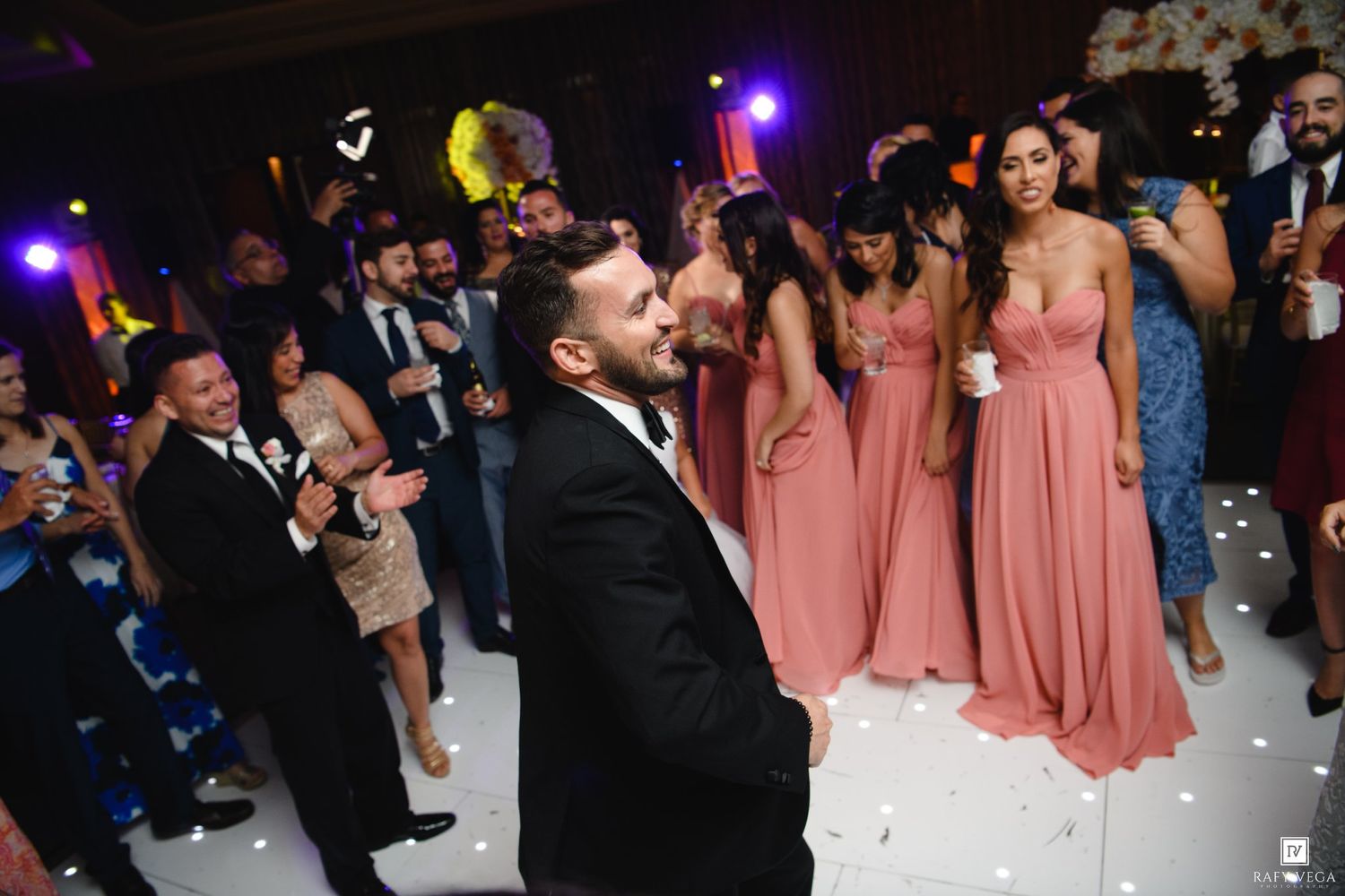 Groom kissing bride on dance floor surrounded by bridesmaids in pink dresses and wedding guests at reception.