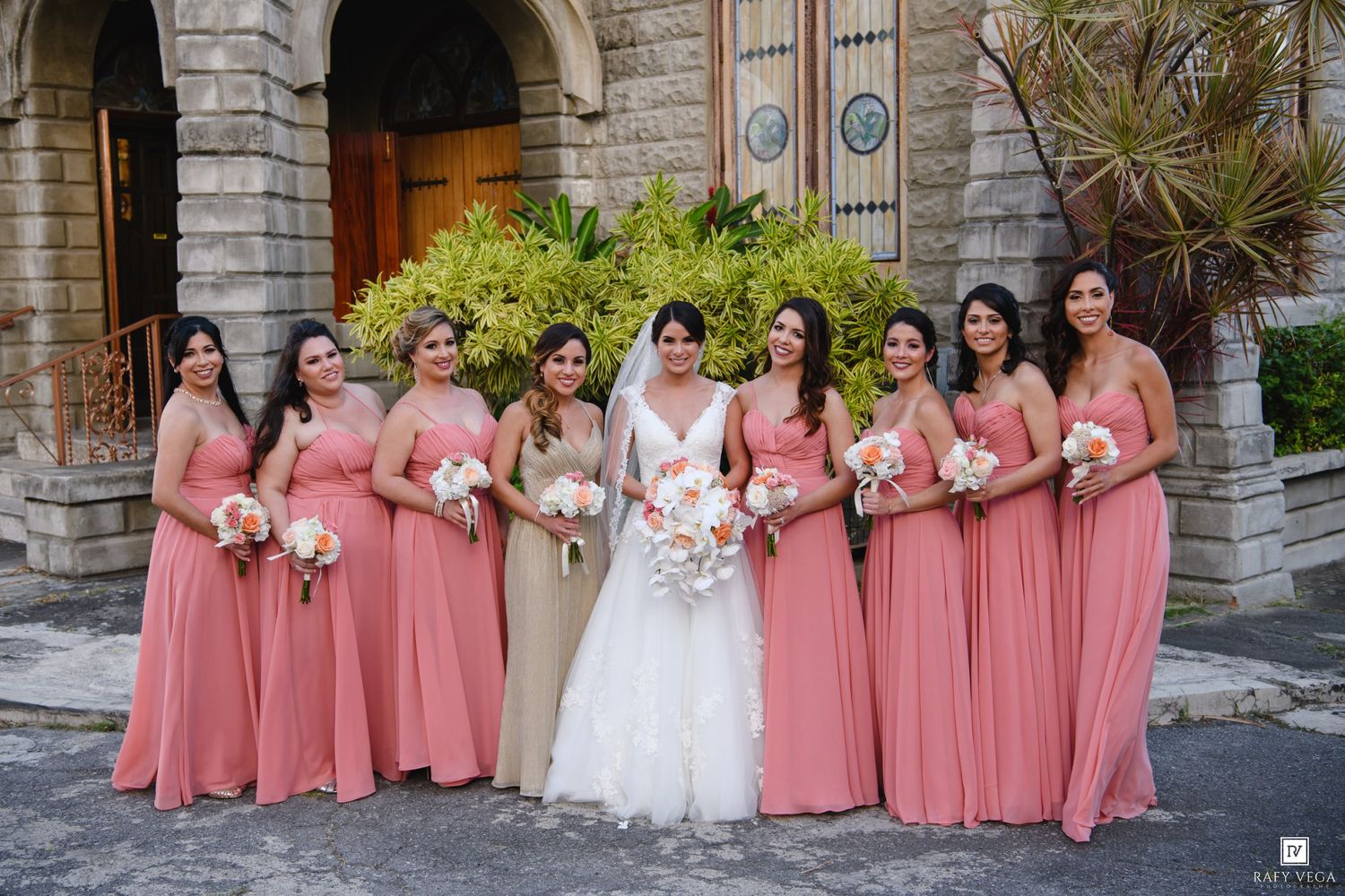 Bride poses with eight bridesmaids in coral pink gowns holding bouquets outside a stone church.