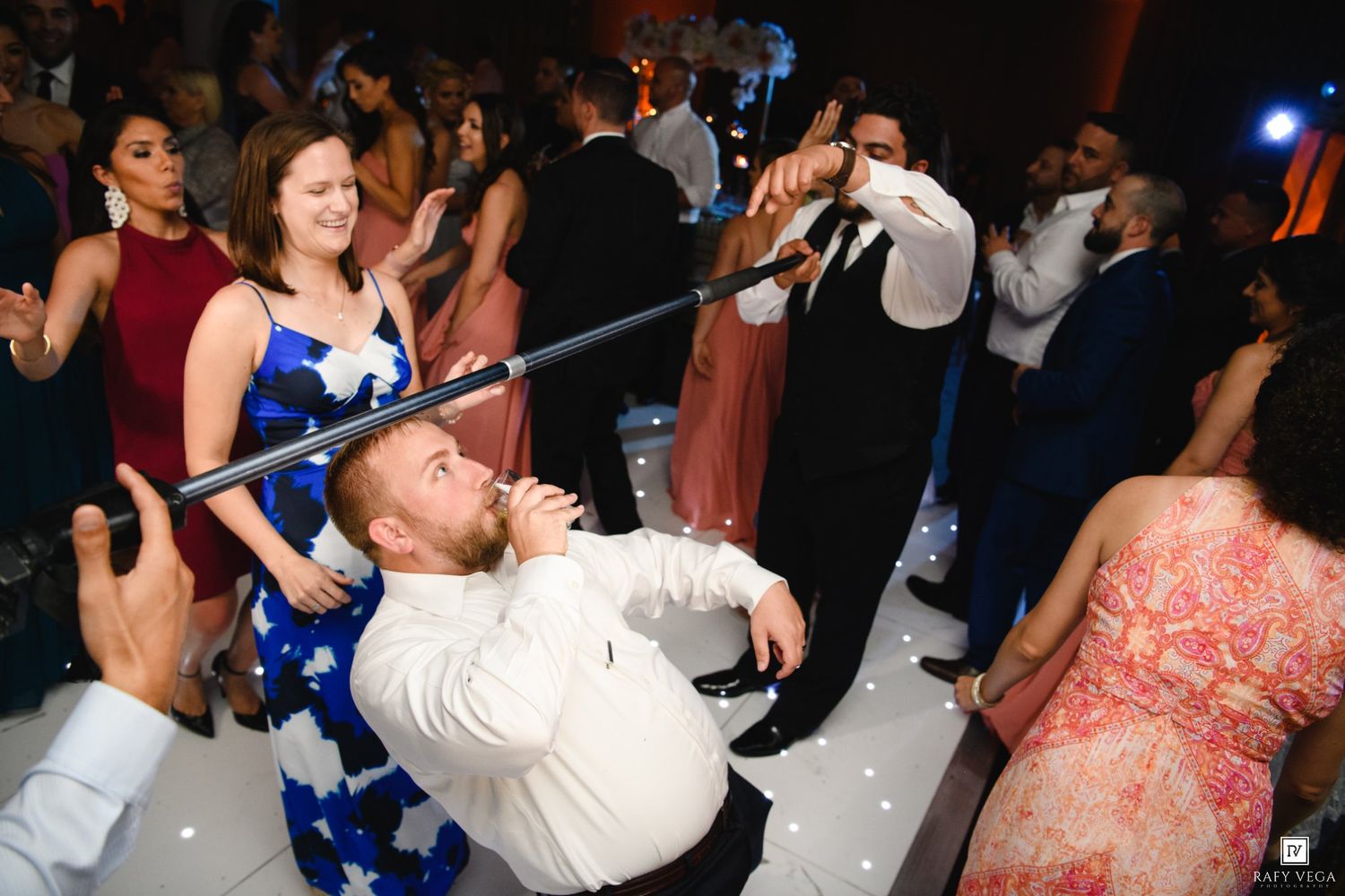 Guest does limbo dance at lively wedding reception party on white dance floor.