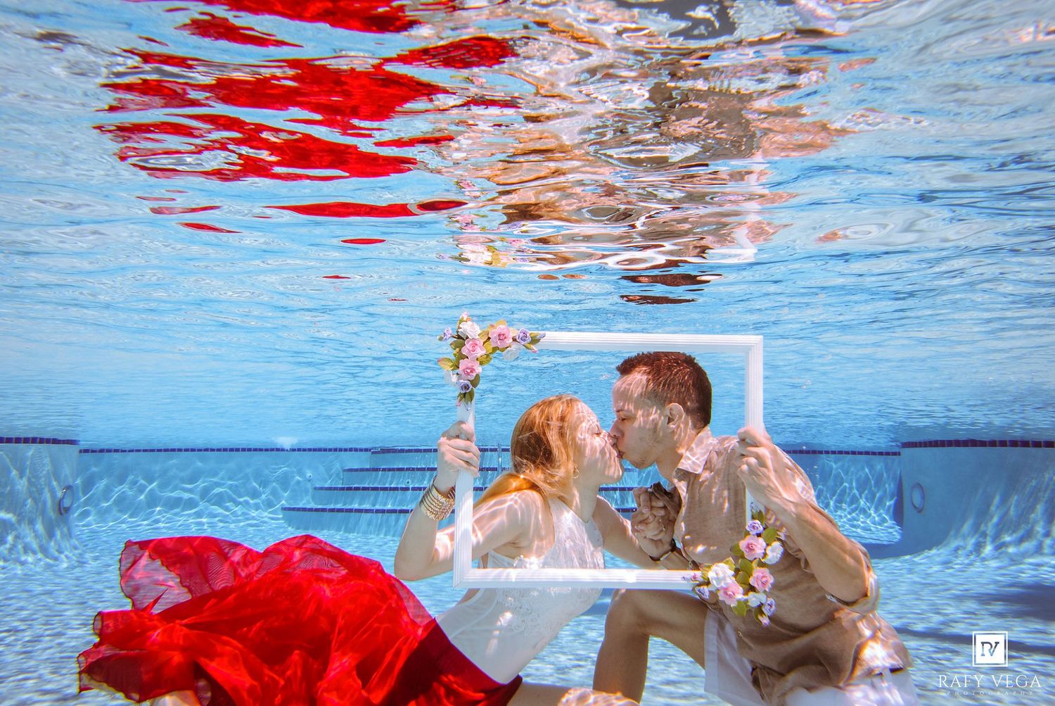 Couple kissing underwater in a pool, holding a white frame with flowers, woman in red dress, romantic underwater wedding photo.