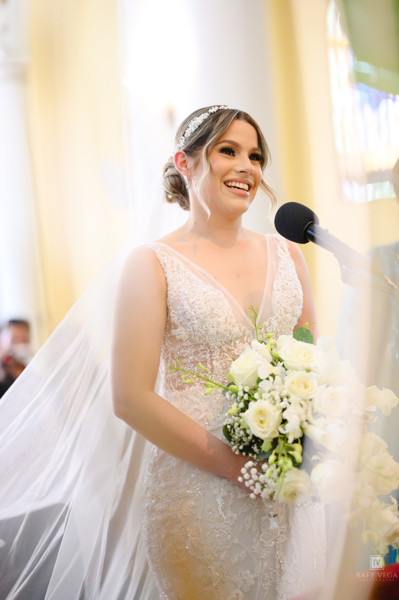Smiling bride in beaded wedding gown holding white bouquet during church ceremony with veil and tiara.