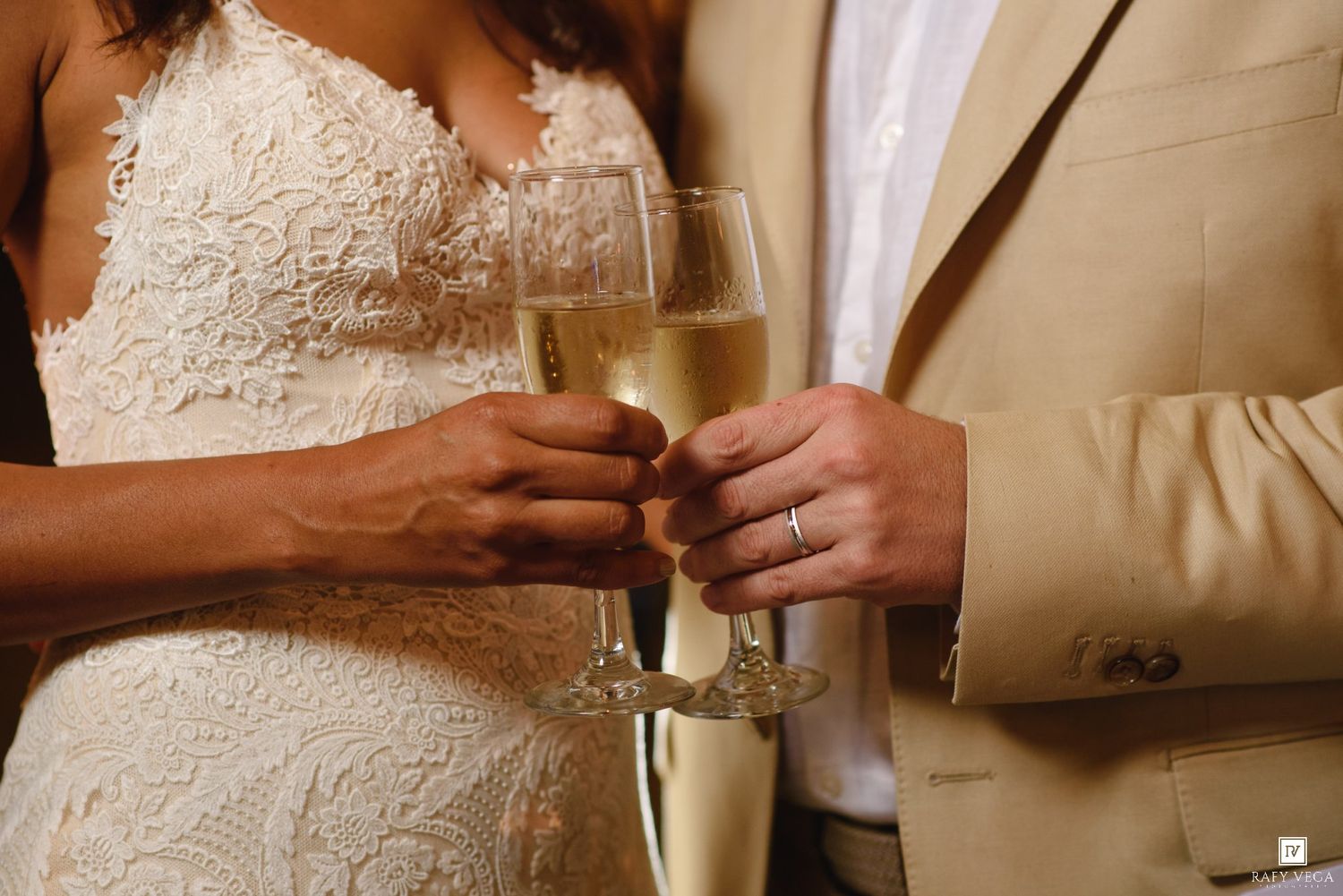 Bride and groom clinking champagne glasses during wedding toast, wearing white lace gown and tan suit.