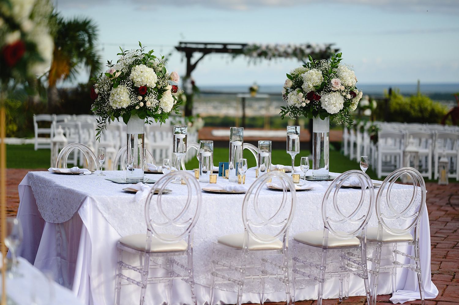 Elegant outdoor wedding reception table with white linens, crystal chairs, and floral centerpieces overlooking the ocean.