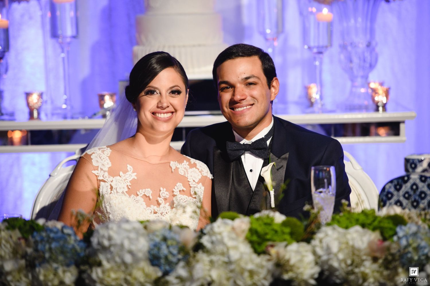 Bride and groom smiling at wedding reception sweetheart table with floral arrangements and candlelit backdrop.