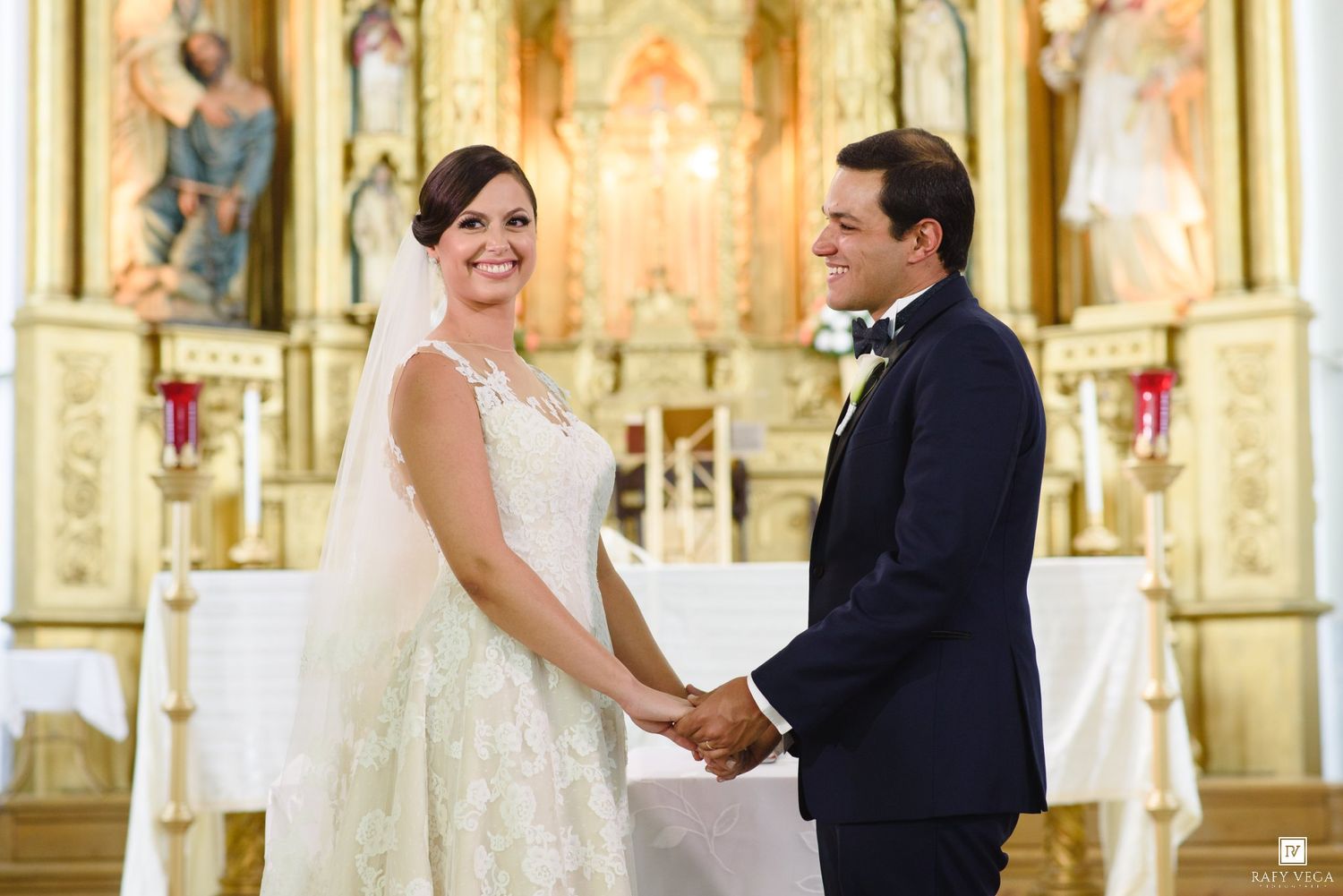 Bride and groom holding hands and smiling at the altar of a beautiful golden church during their wedding ceremony.