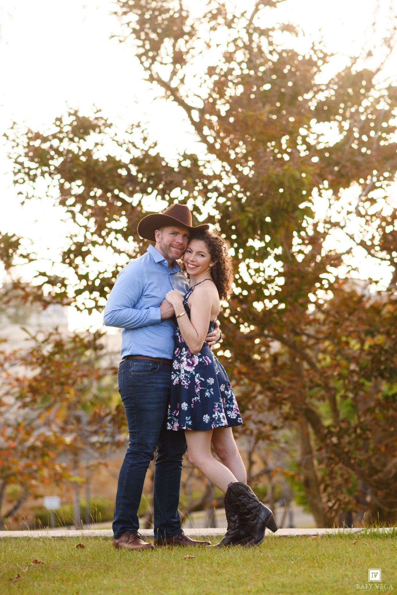 Couple in cowboy hats embracing outdoors, woman in floral dress and boots, man in blue shirt, golden sunlit tree backdrop.