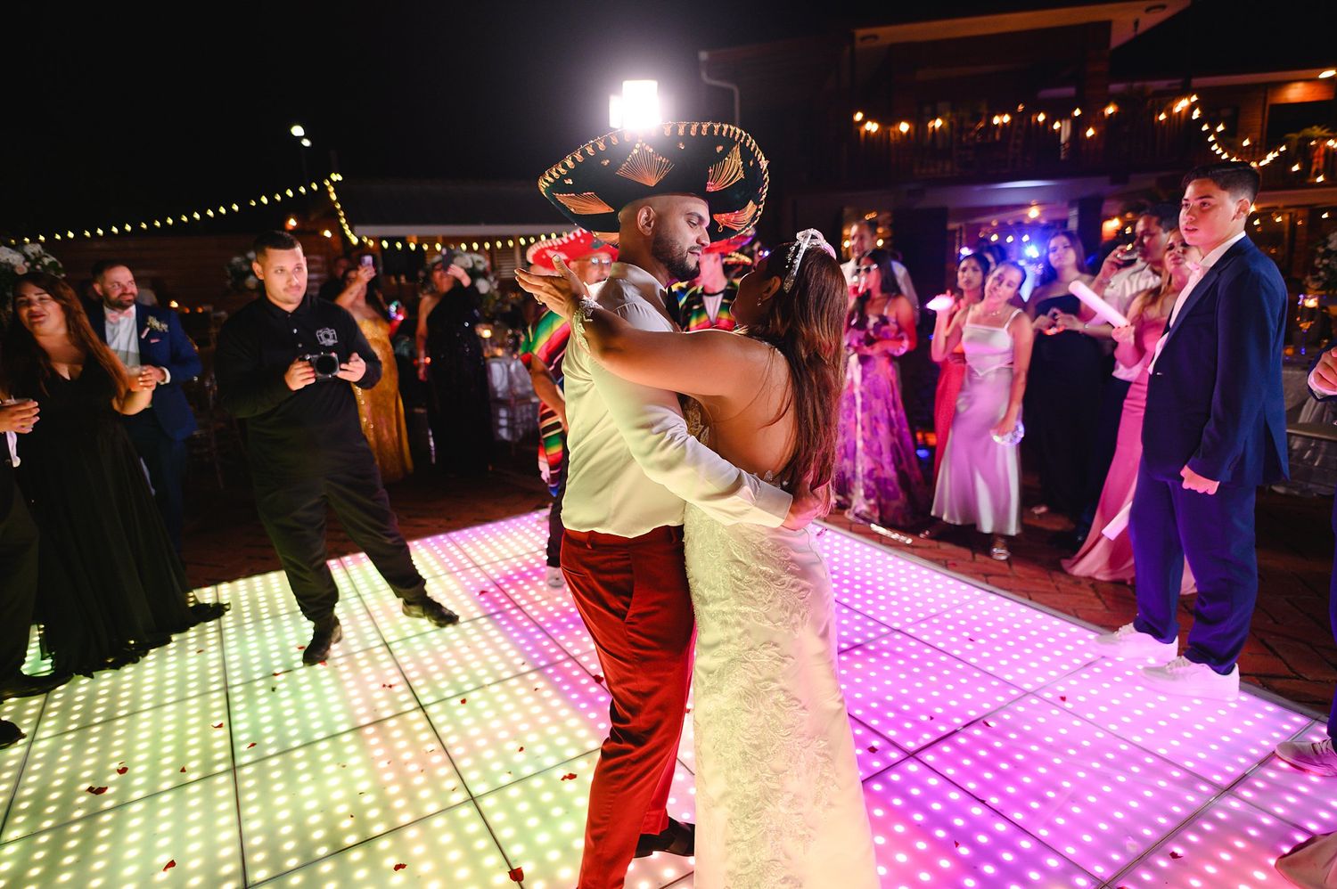 Wedding guests dance on illuminated LED floor as mariachi performer joins celebration at night.