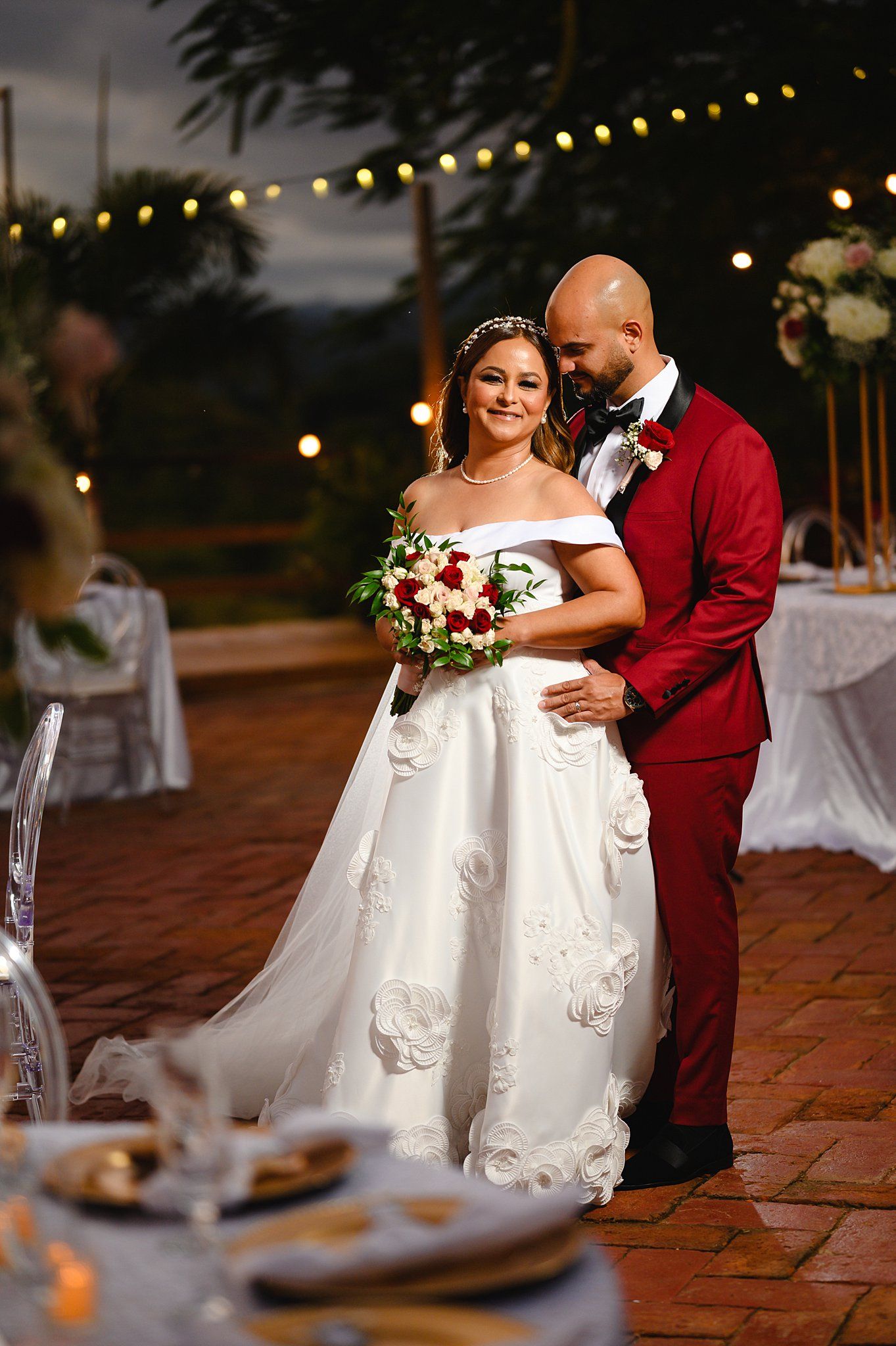 Bride in white off-shoulder gown holds red bouquet beside groom in red suit at outdoor evening wedding reception.