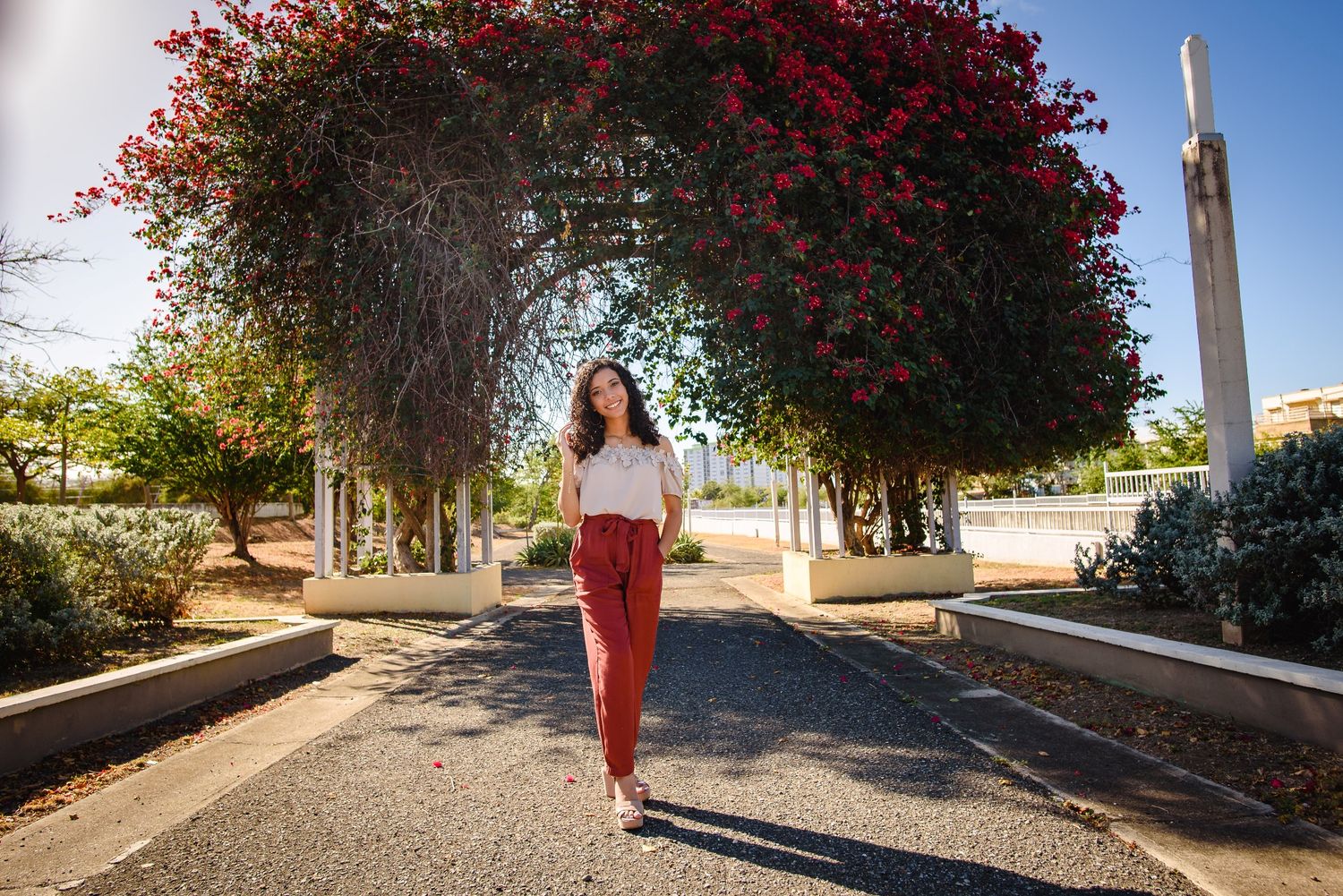 Woman in red pants walks under a vibrant bougainvillea arch on a sunny garden pathway.