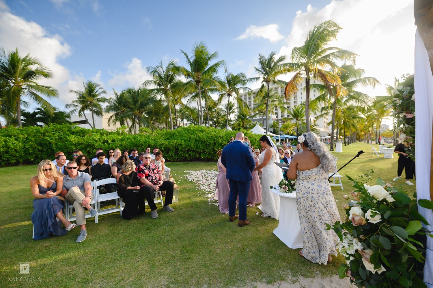 Outdoor wedding ceremony on tropical lawn with palm trees, guests seated on white benches, bride and groom exchanging vows.