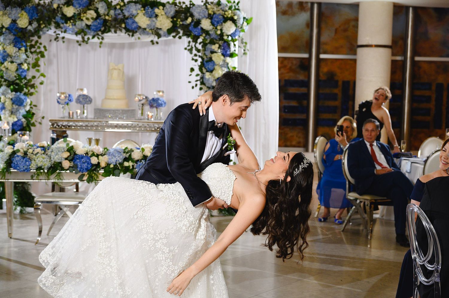 Bride and groom share romantic dip dance at elegant wedding reception with blue floral arch and white cake backdrop.