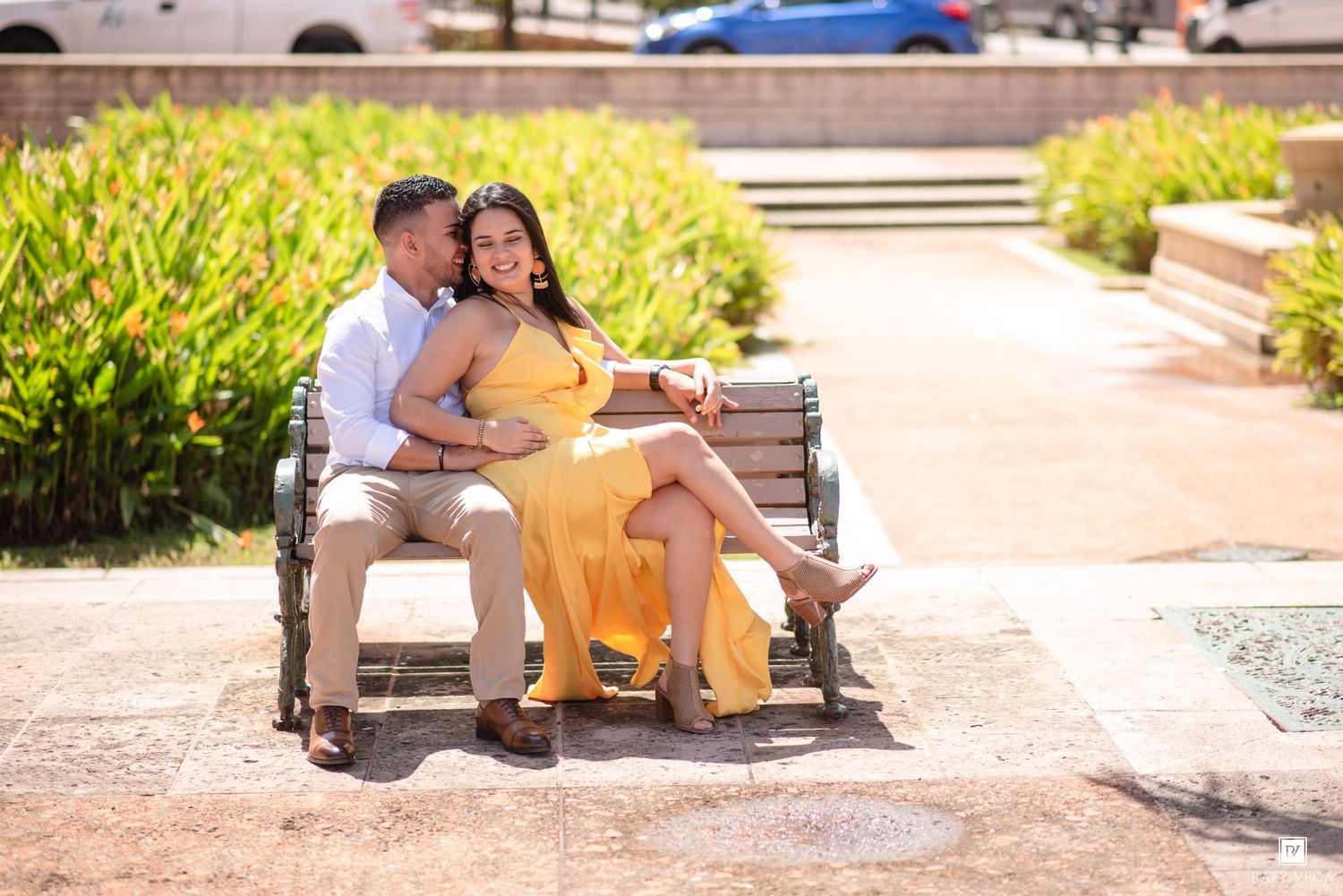Couple sitting together on a park bench, woman in yellow dress, man in white shirt, sunny outdoor setting.