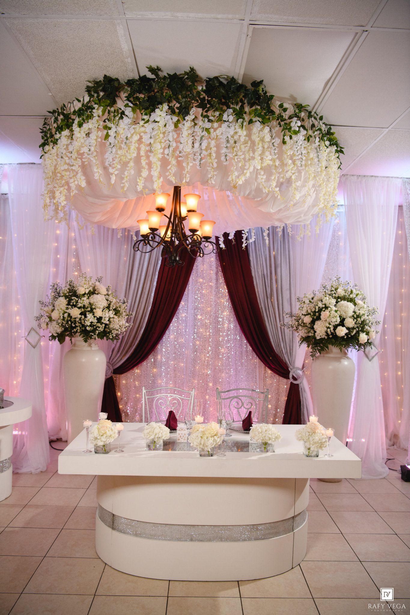 Elegant wedding sweetheart table with floral chandelier, burgundy drapes, white roses, and candlelight in a decorated reception hall.