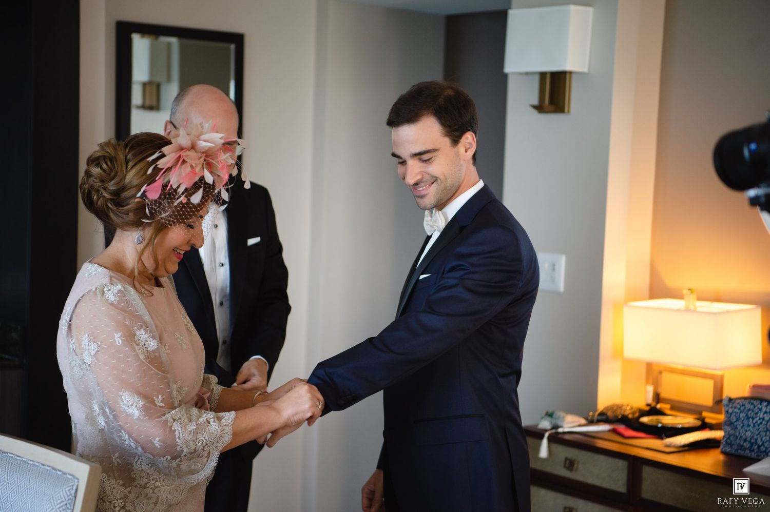 Groom in navy suit shaking hands with bride in floral dress during wedding preparations indoors.