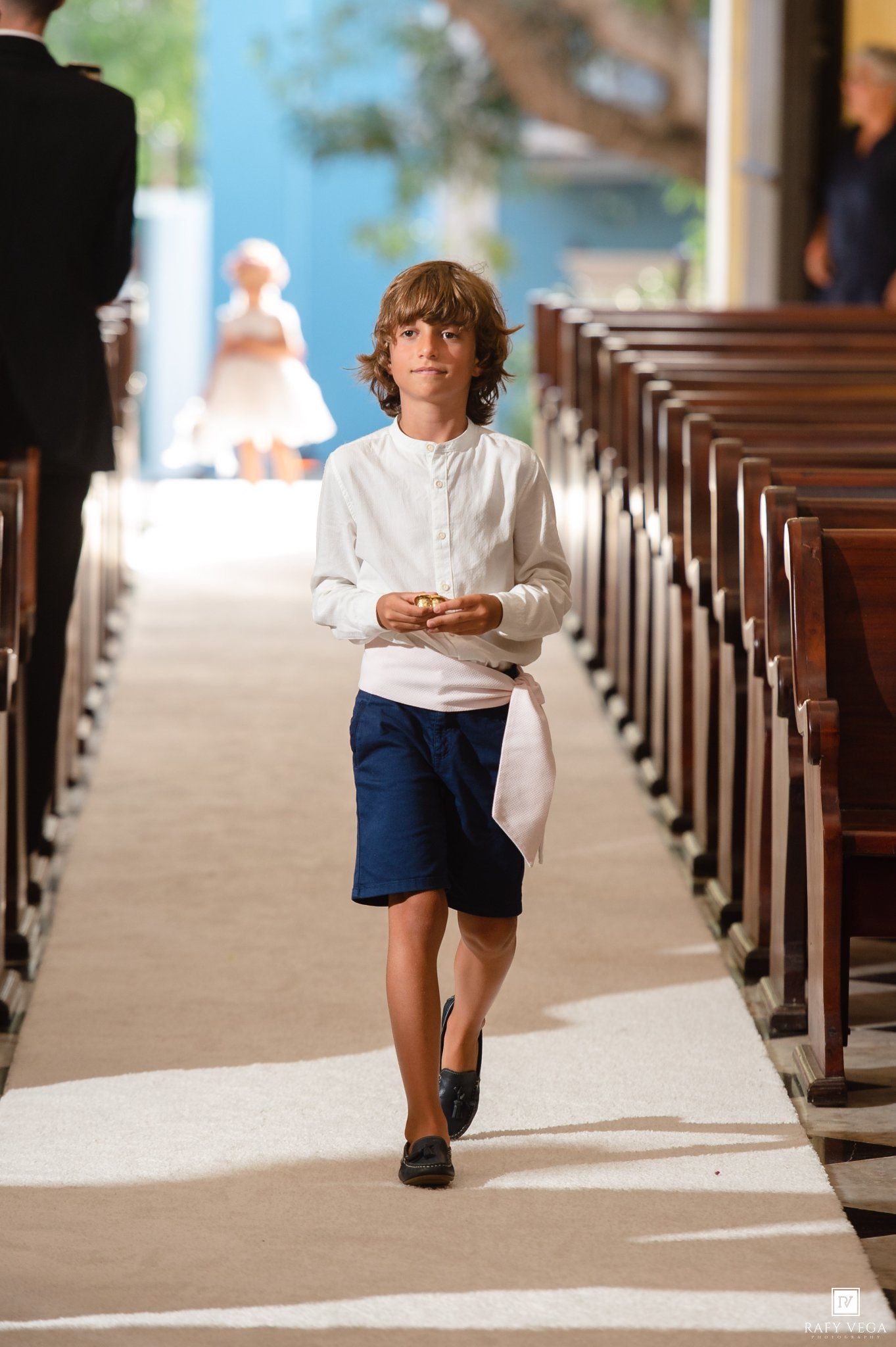 A young ring bearer walks down the church aisle in blue shorts and white shirt during a wedding ceremony.
