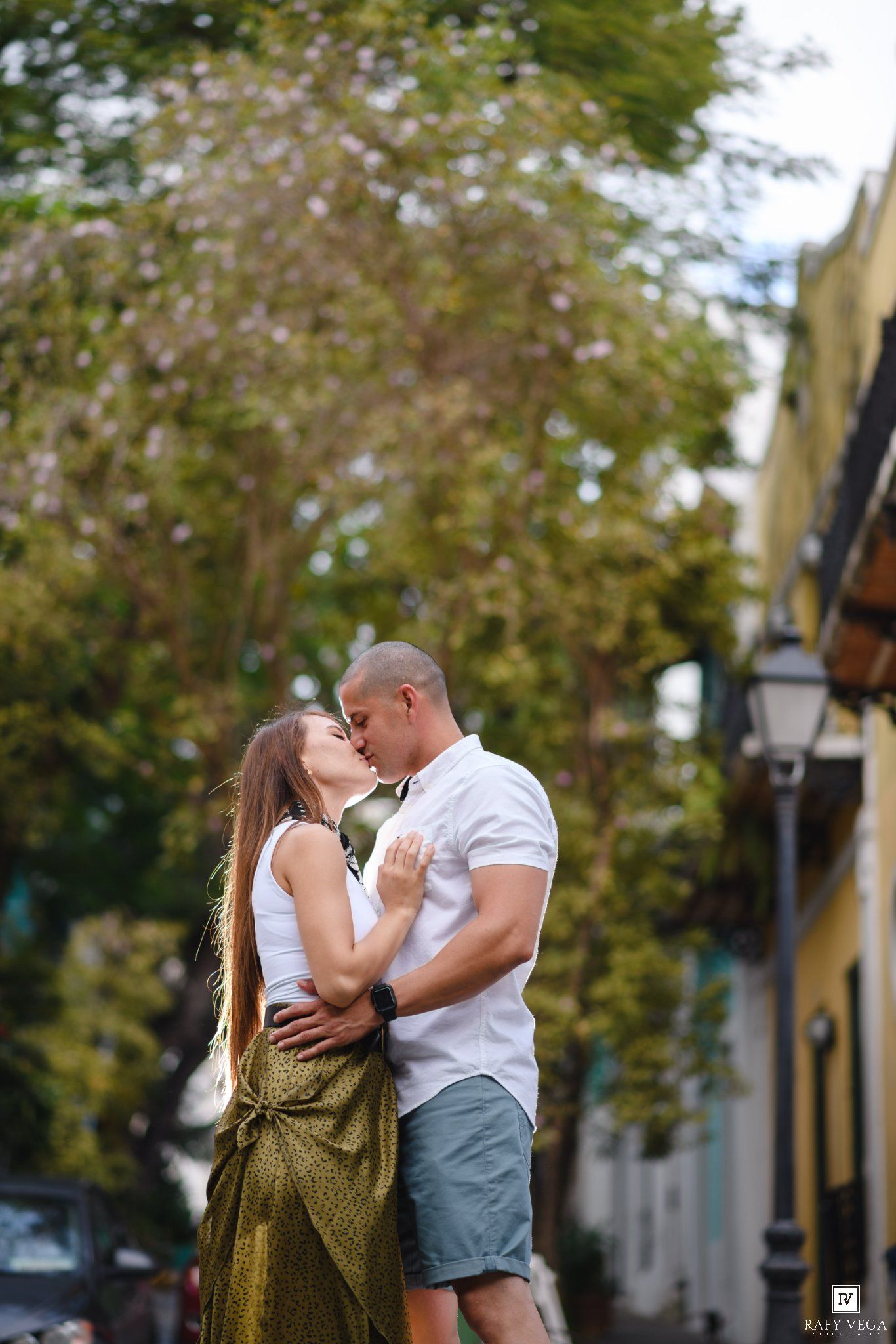 Couple sharing a romantic kiss outdoors on a colorful street, surrounded by lush green trees and warm natural light.