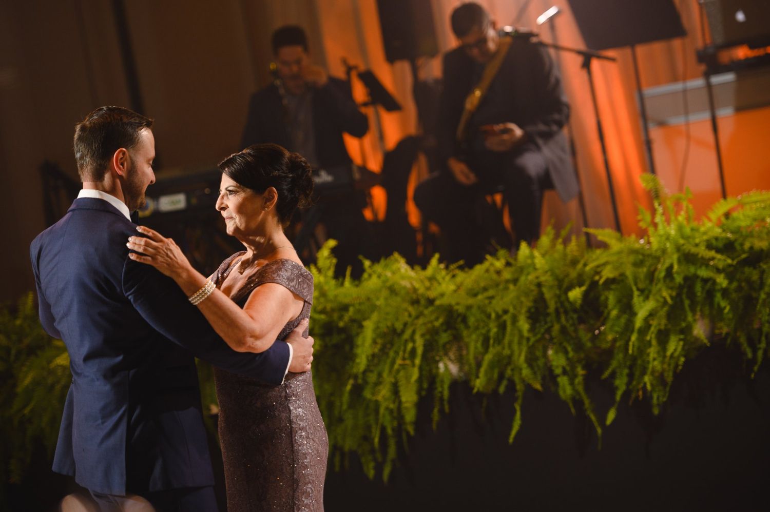 Couple sharing a romantic dance at an elegant wedding reception with live band and lush green ferns in background.