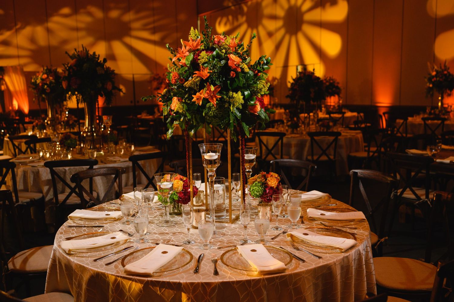 Elegant wedding reception with gold tablecloths, tall floral centerpieces, and warm amber lighting in a ballroom.
