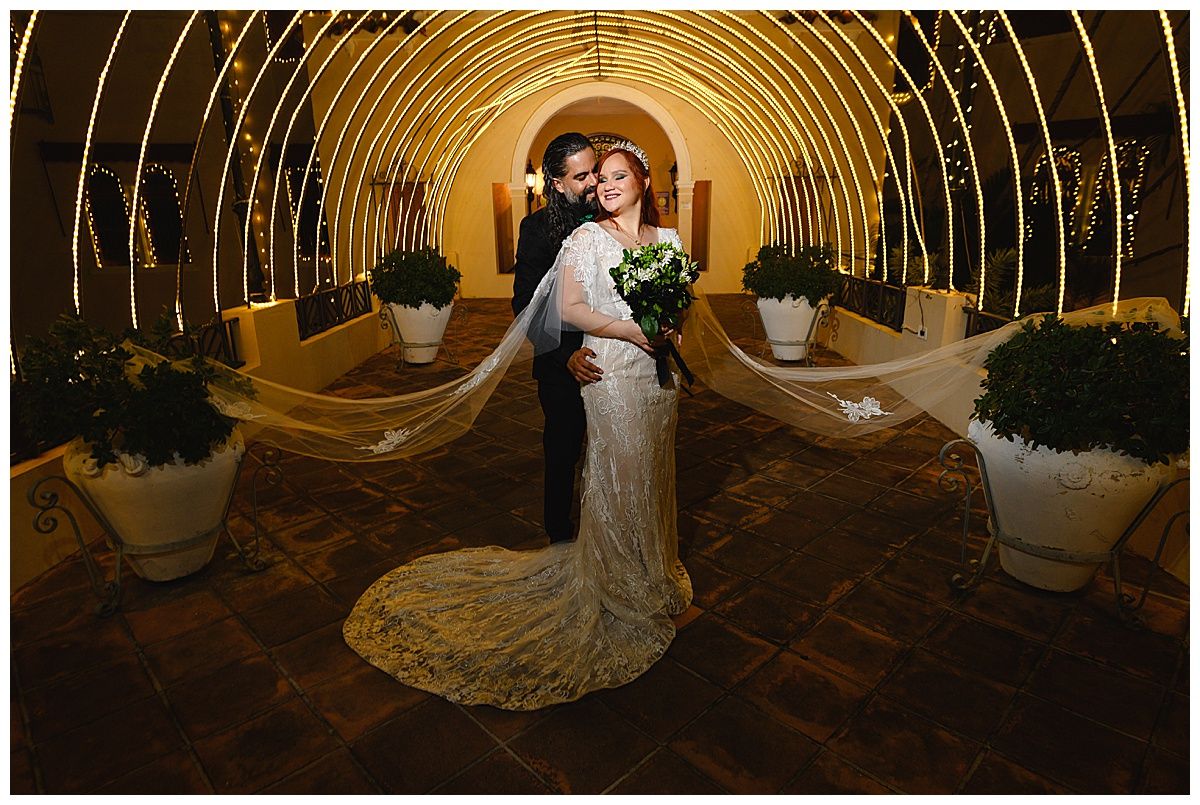 Bride and groom pose romantically under glowing arch tunnel with flowing veil at elegant wedding venue.