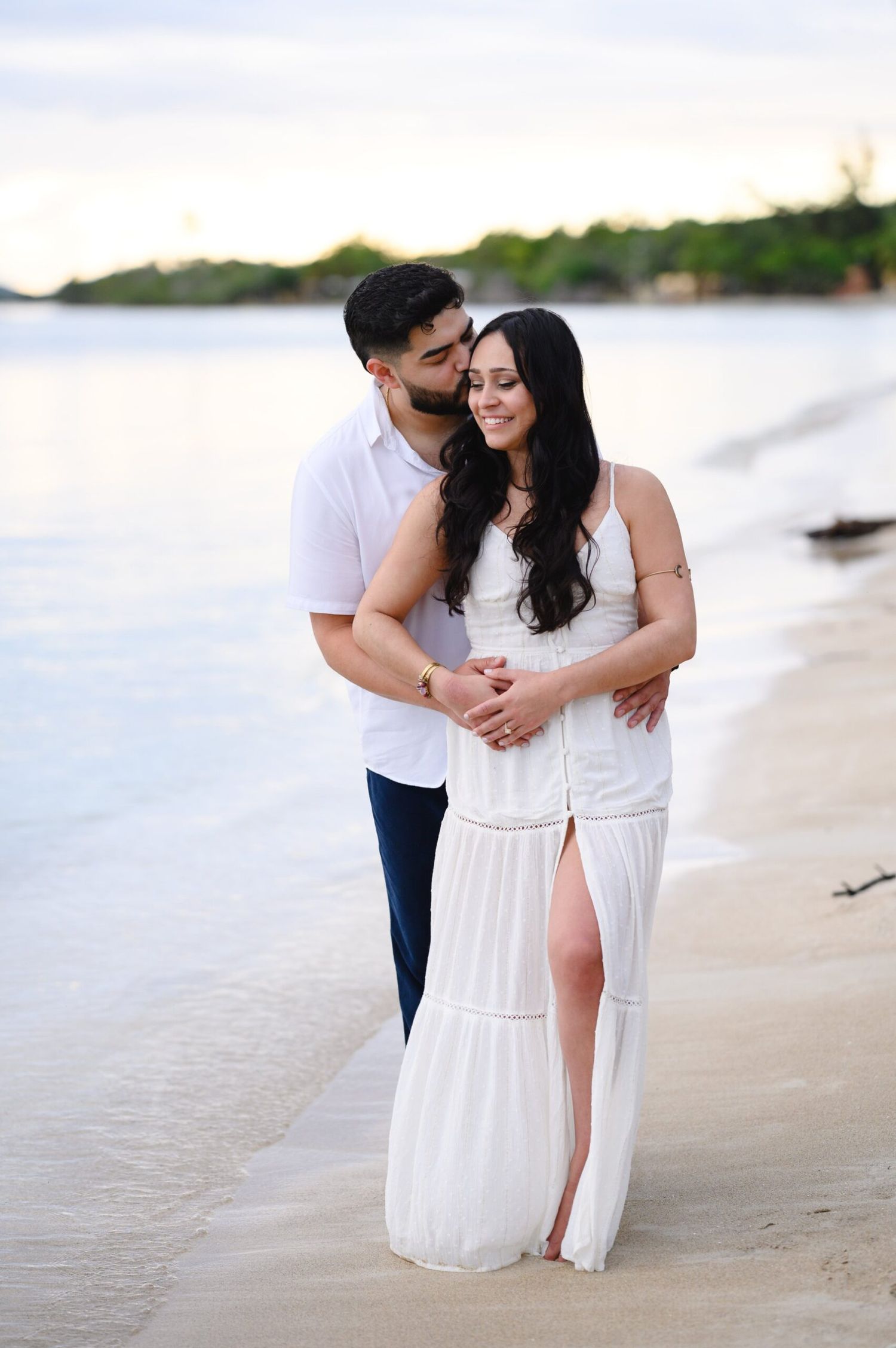 Man kissing woman on cheek while embracing on beach at sunset, couple in white and navy outfits.
