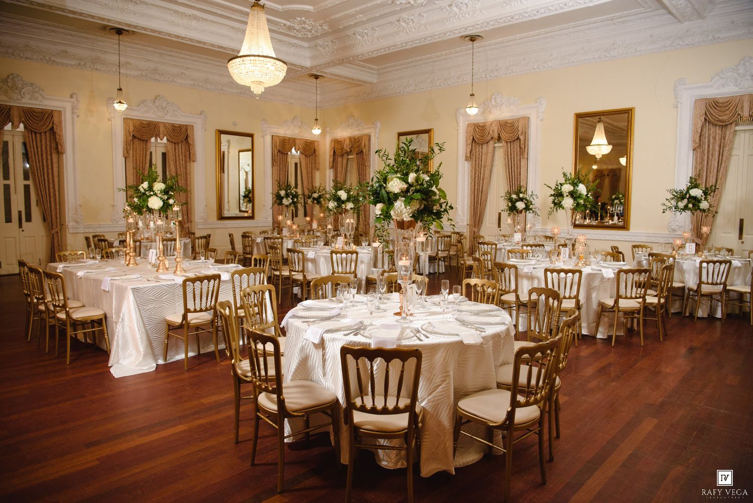 Grand wedding reception hall with gold Chiavari chairs, white round tables, tall floral centerpieces, and hardwood floors.