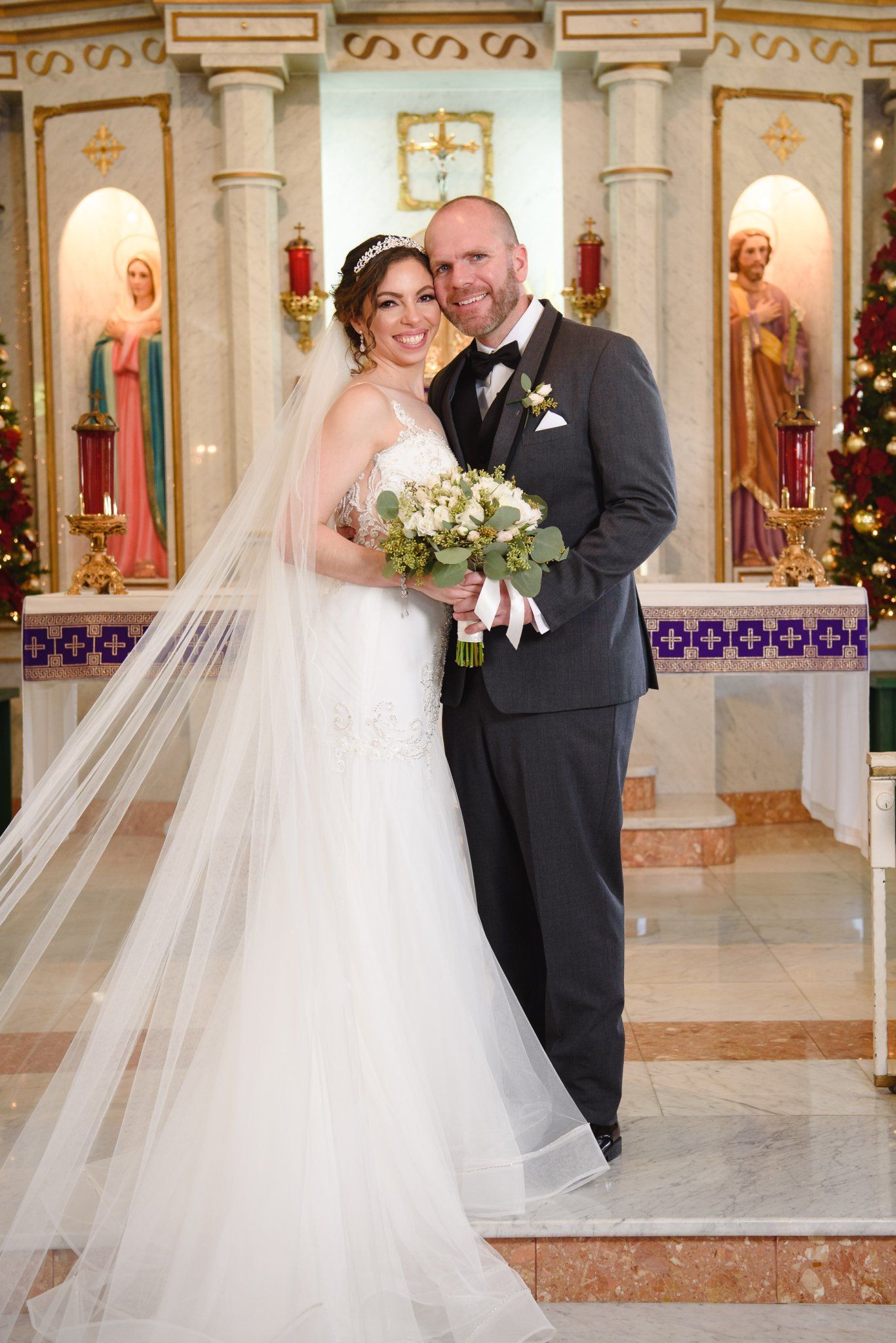 Bride and groom pose together at church altar, bride in white gown with veil holding bouquet, groom in gray suit.