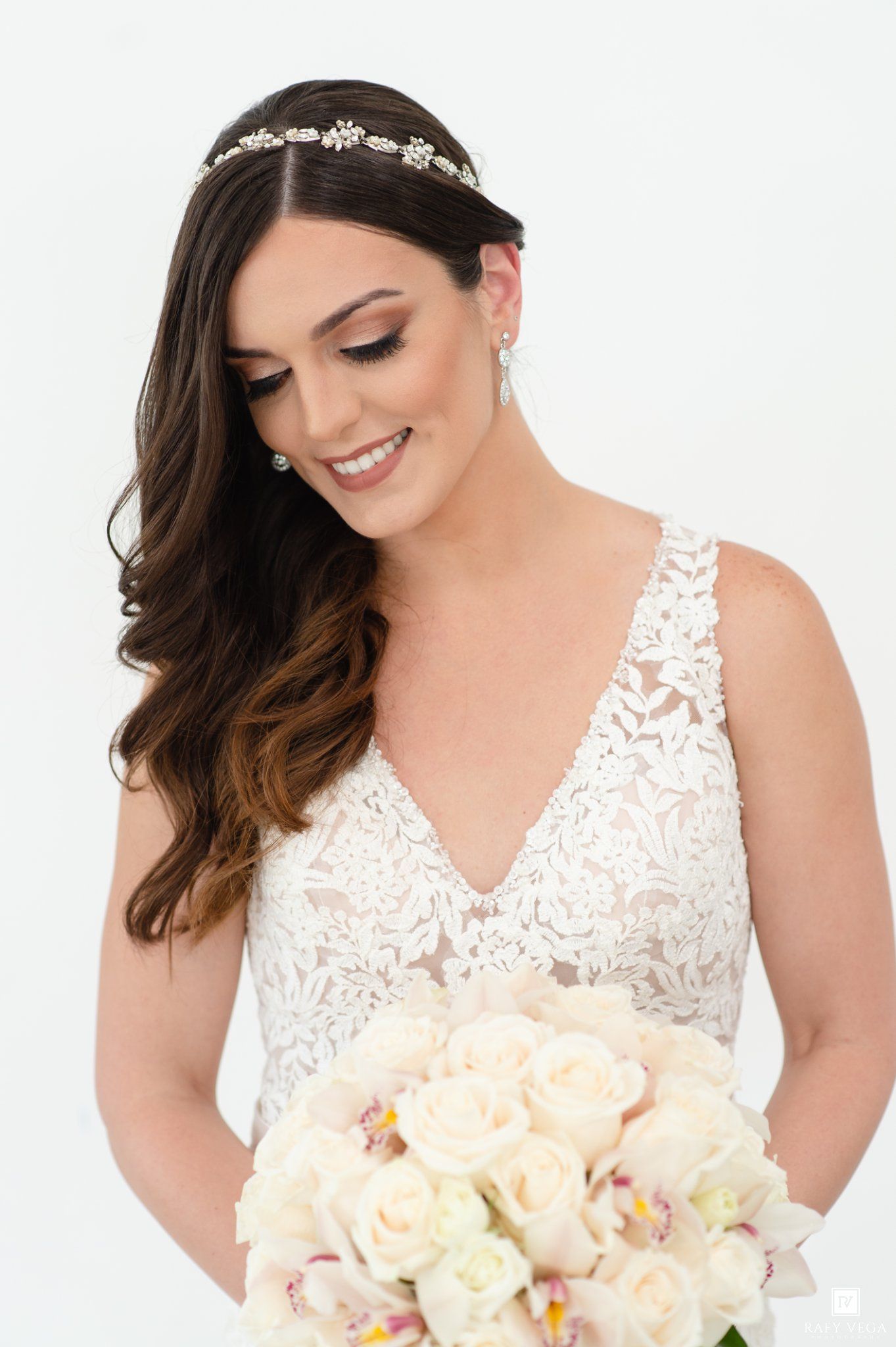 Bride in lace V-neck gown with pearl headband looks down at white rose bouquet.