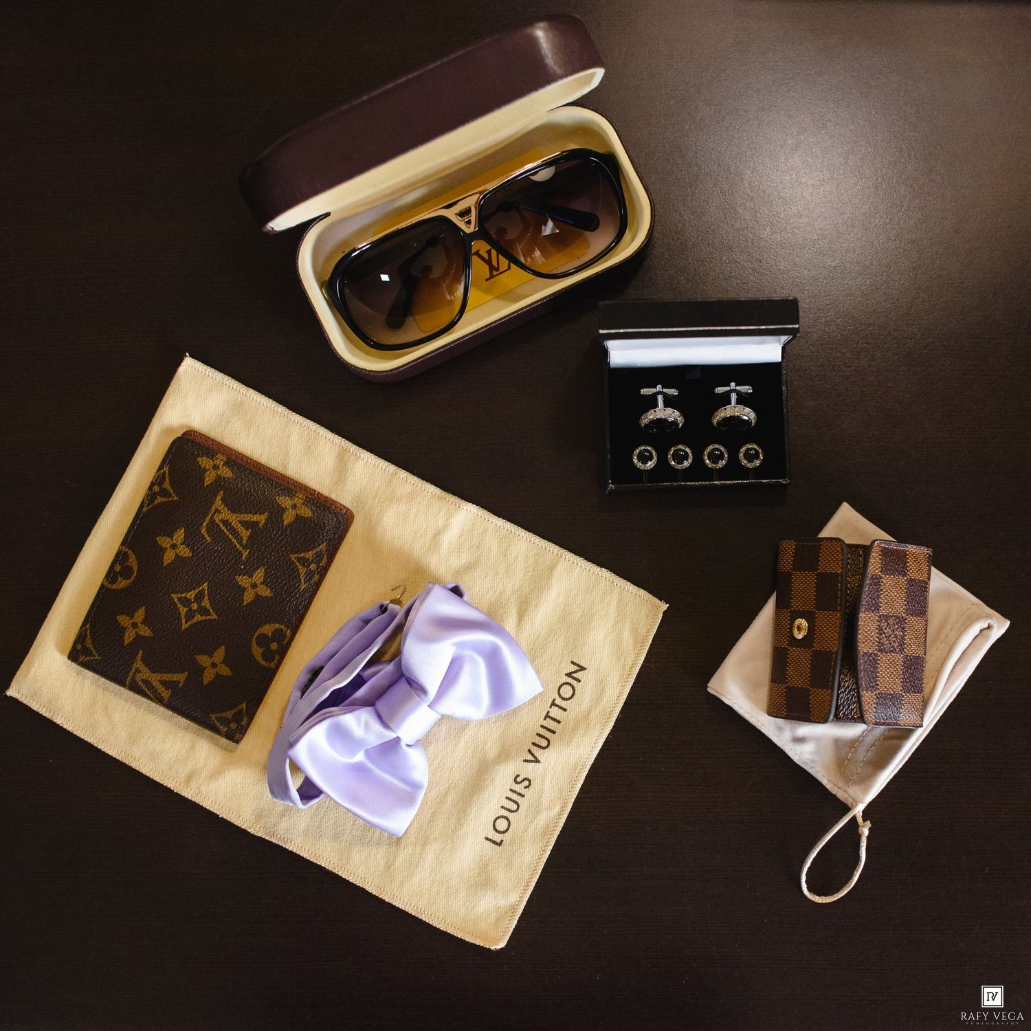 Luxury Louis Vuitton accessories including wallet, key holder, sunglasses, cufflinks, and dust bag arranged on dark surface.