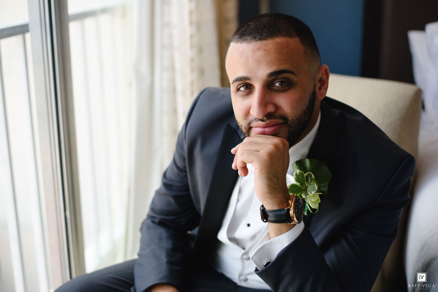 Groom in navy suit with boutonniere sitting thoughtfully by window on wedding day.