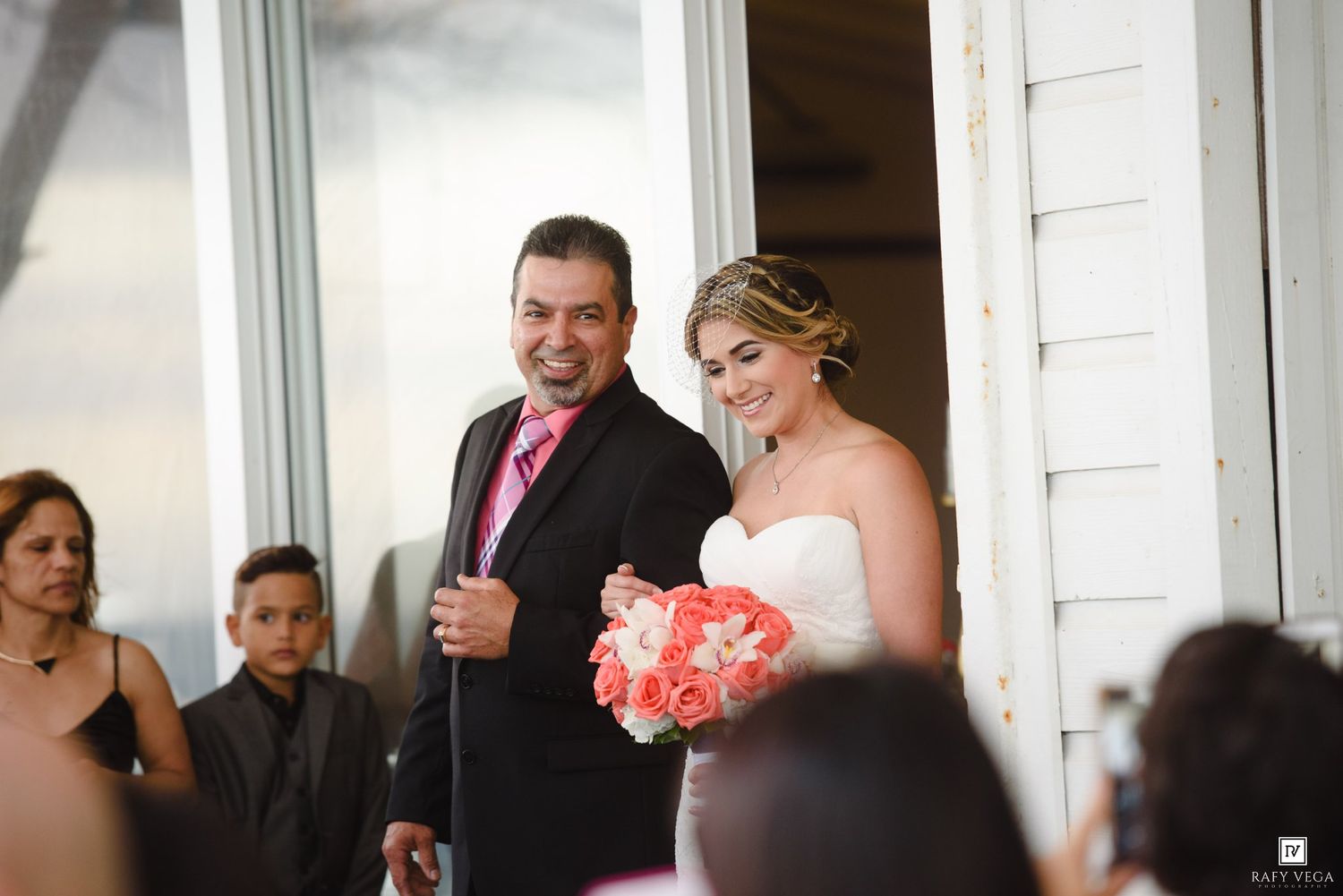 Smiling bride holding coral bouquet stands with groom in black suit during wedding ceremony entrance.