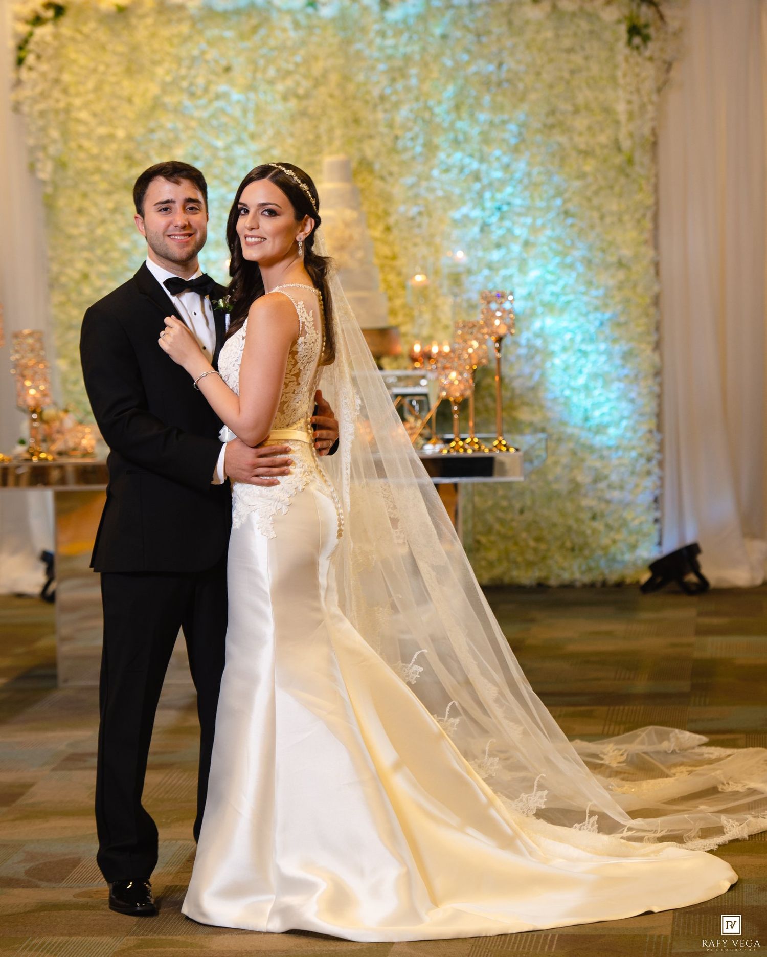 Elegant bride and groom posing together at wedding reception with floral wall backdrop and candlelit cake table.