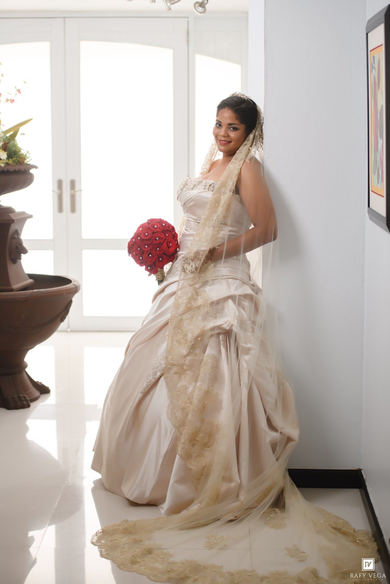 Bride in champagne gold lace wedding gown holding red bouquet, posing elegantly by white wall with veil.