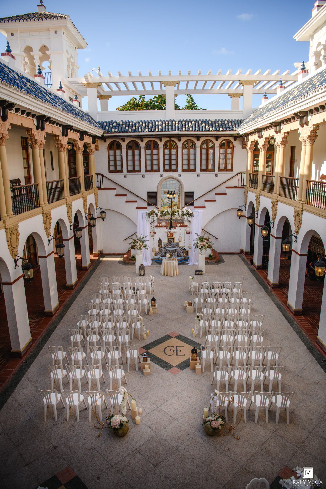 Elegant outdoor wedding ceremony setup in a white Spanish courtyard with rows of white chairs and floral decorations.