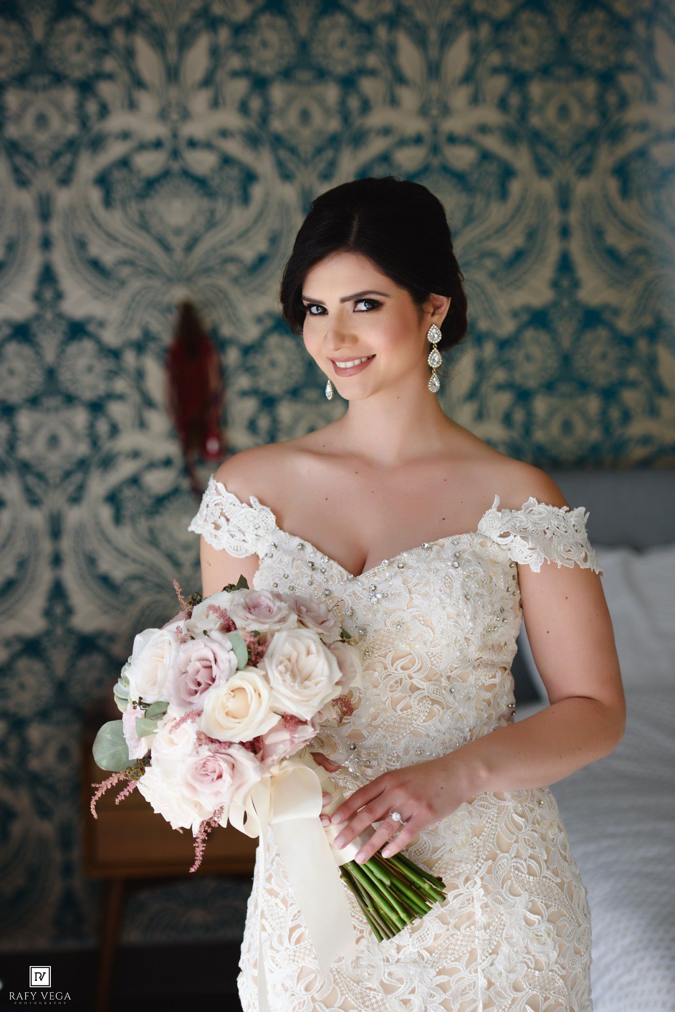 Bride in off-shoulder lace wedding gown holding blush pink rose bouquet, smiling elegantly before floral wallpaper backdrop.