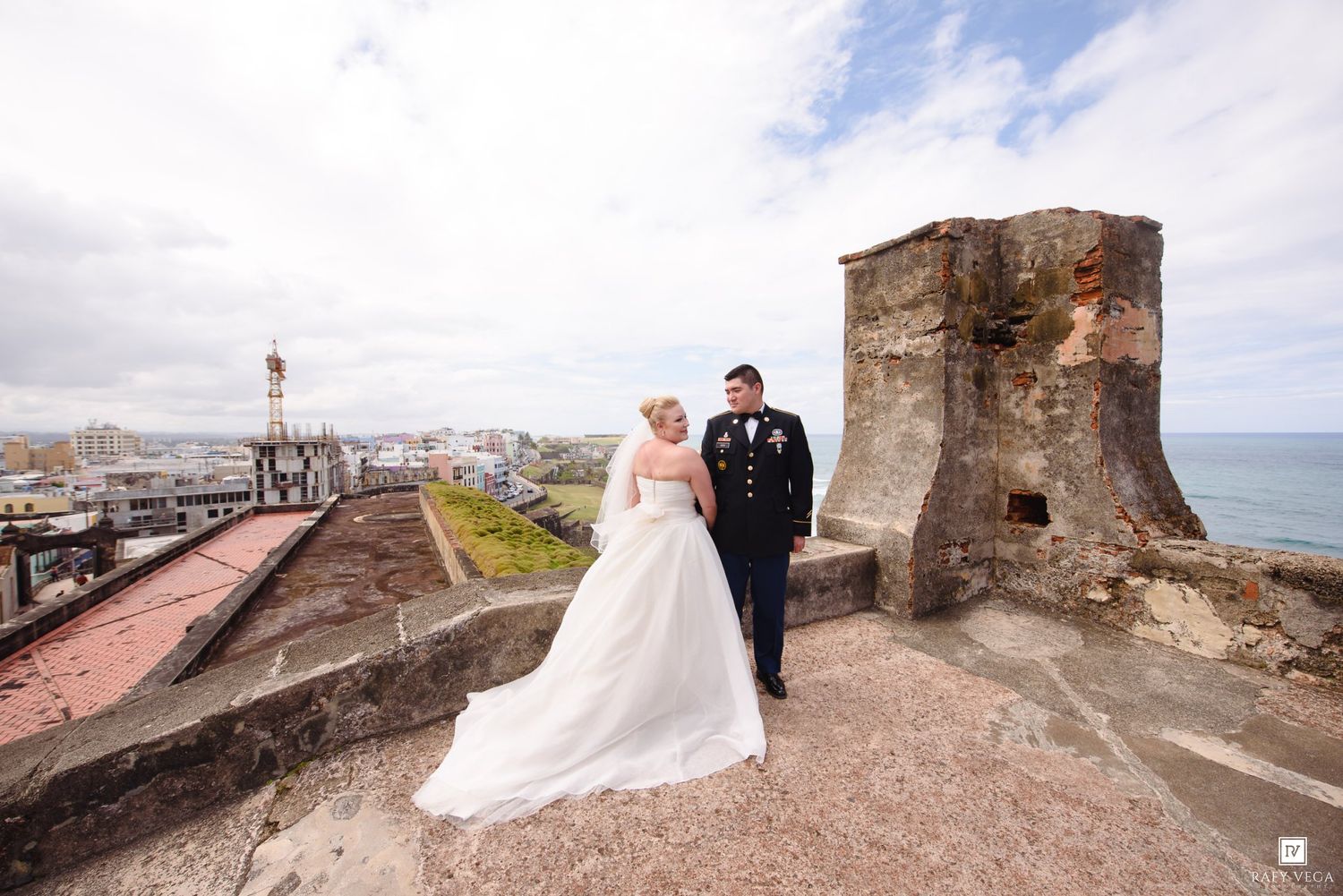 Bride in white gown and military groom pose on historic fort rooftop with coastal city views behind them.