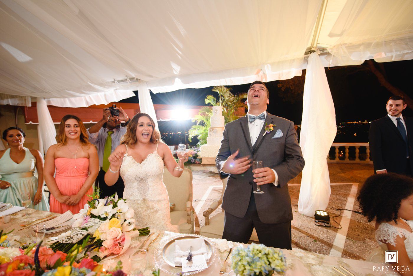 Bride and groom laughing joyfully during wedding reception toast under elegant tent venue.