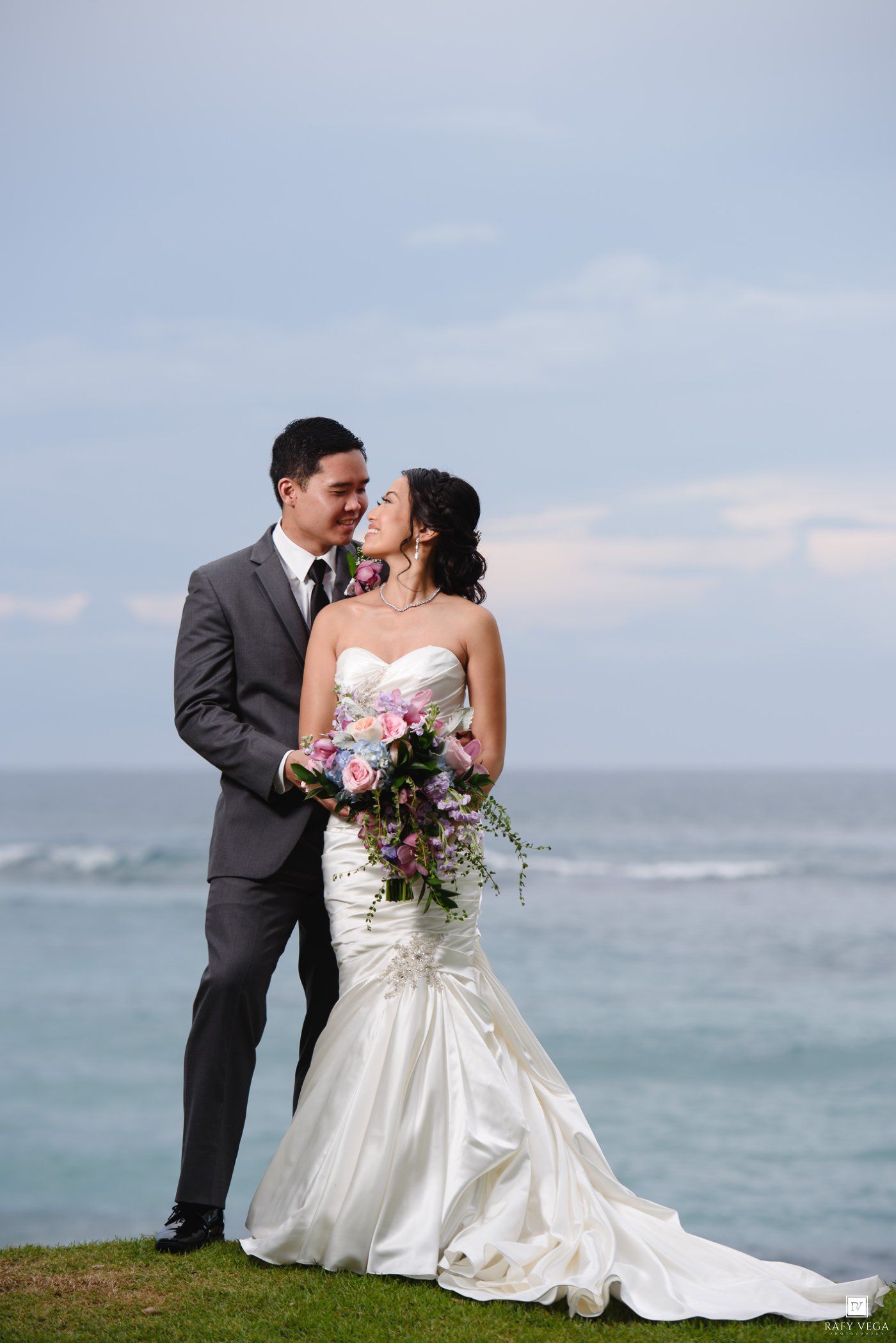 Bride and groom share a romantic moment by the ocean, she in a white mermaid gown holding a bouquet, he in a gray suit.