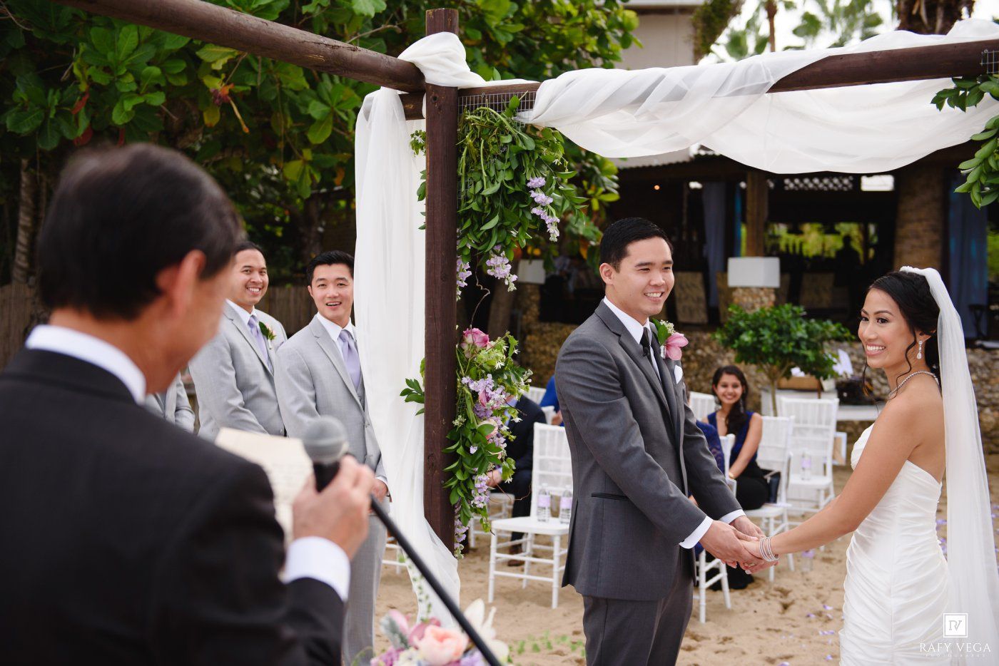 Groom in gray suit holds bride's hands at outdoor beach wedding ceremony beneath floral arch.