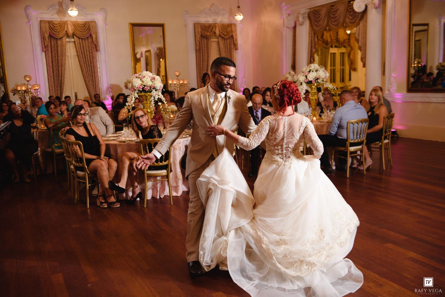Bride and groom share first dance at elegant wedding reception with floral centerpieces and guests seated in background.