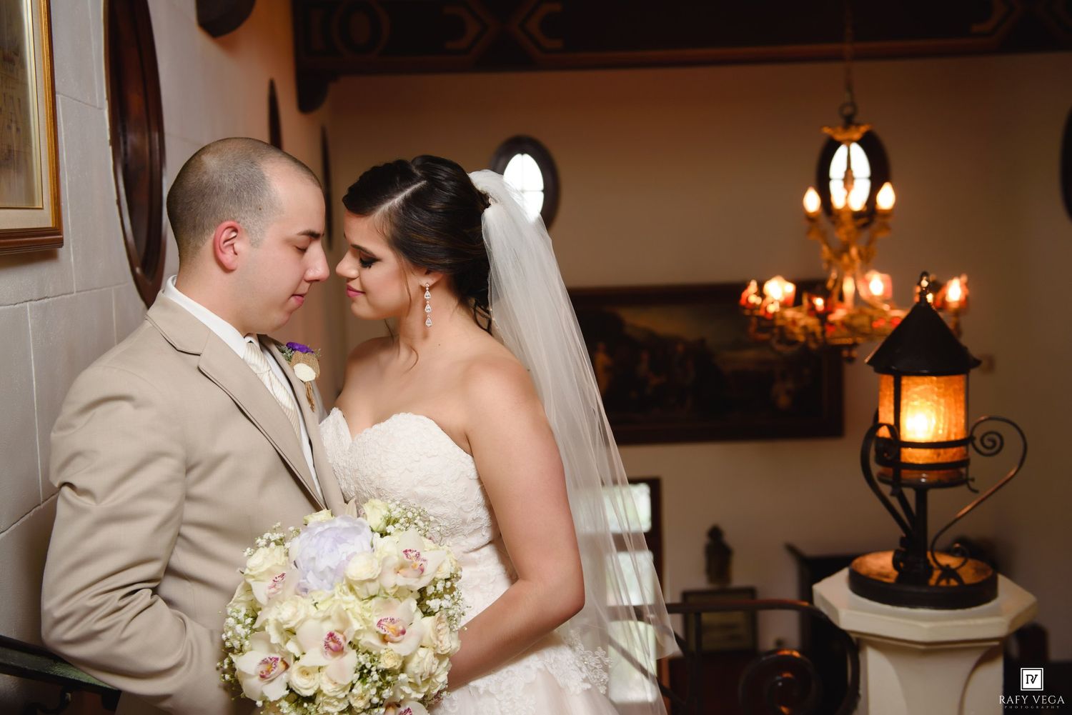 Bride holding white bouquet and groom in tan suit share intimate moment near vintage lantern in rustic venue.