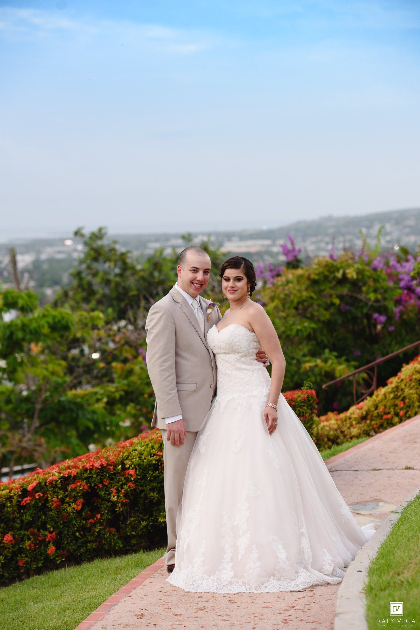 Bride in white lace gown and groom in tan suit pose on garden path with colorful flowers and city view behind them.