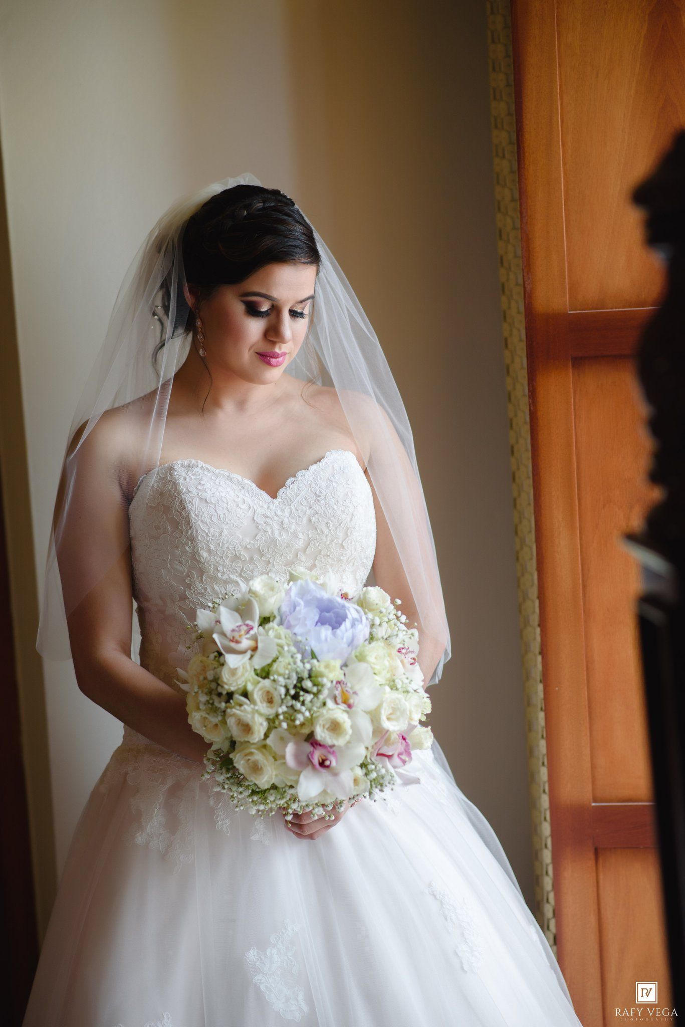 Bride in strapless lace ball gown and veil holds elegant white and pink floral bouquet near a window.