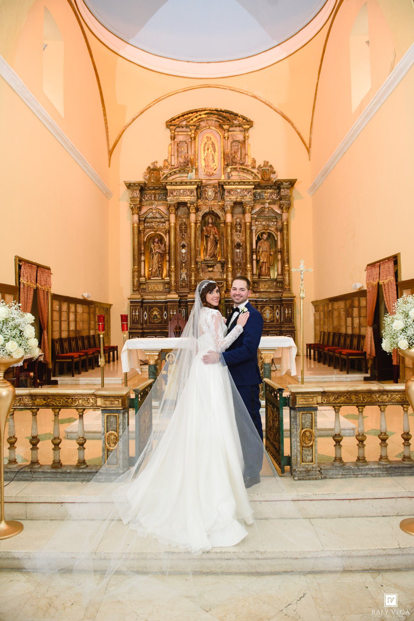 Bride and groom posing together at ornate church altar with golden reredos and white floral arrangements.