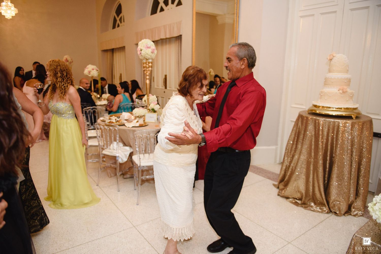 Couple dancing at a wedding reception with guests, tables, and a tiered white wedding cake in the background.