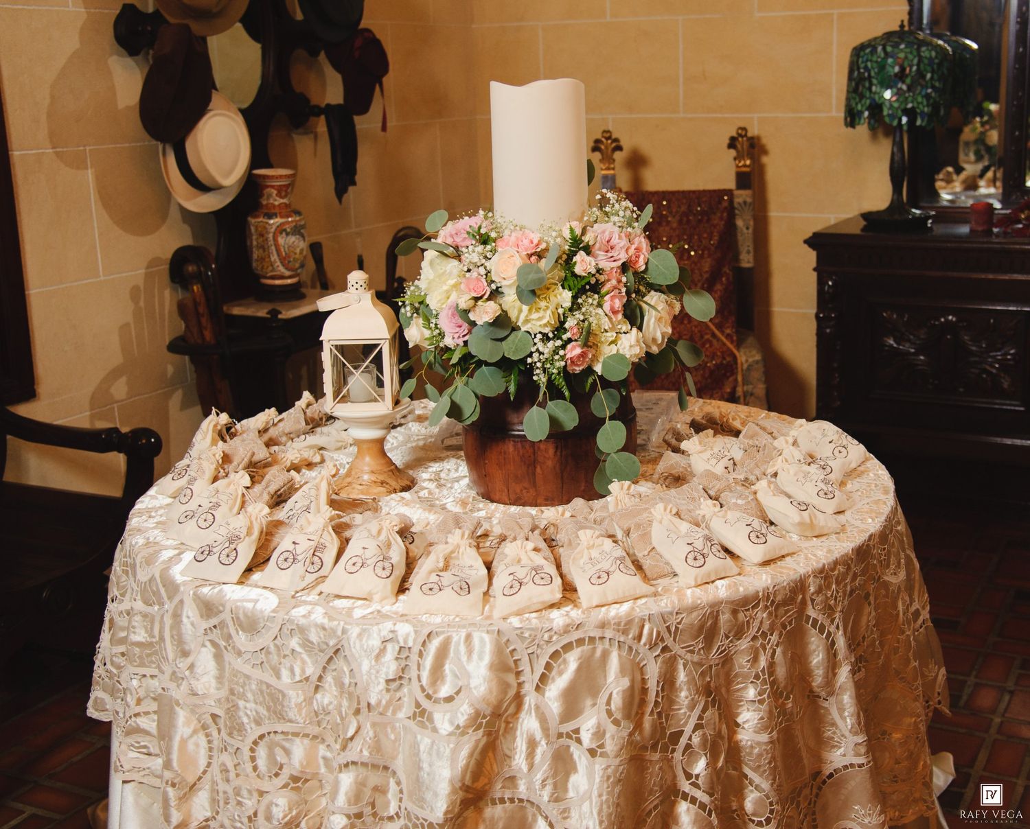 Elegant wedding escort card table with floral centerpiece, white candle, lantern, and gold lace tablecloth.